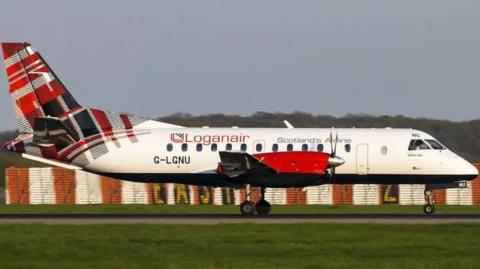 A Loganair plane landing on a runway that is in between two grassy patches. It is predominantly white, the wing is red, and the tail has a red, black and white tartan motif. 