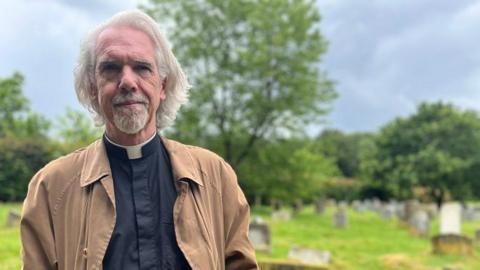 The Reverend Tim Heaney, wearing a clerical collar and a brown coat, stands in a grassy graveyard with scattered headstones, trees and hedges in the background under an overcast sky.