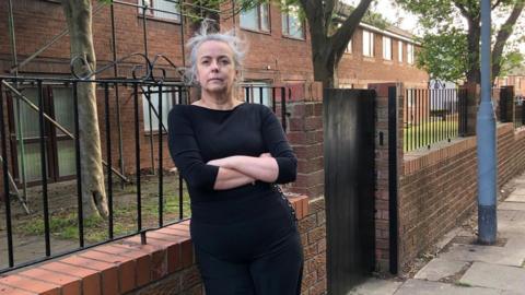 Thelma Clark has grey hair and a black top and black trousers. She is standing with her arms crossed in front of a brick wall. 