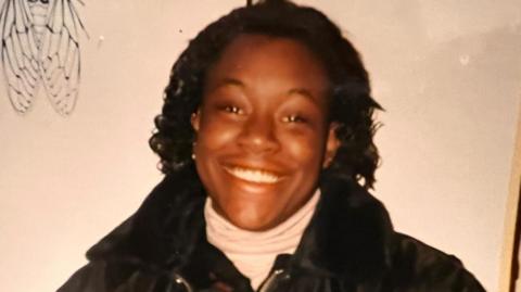A woman with shoulder-length black hair smiling at the camera. She is wearing a black jacket.