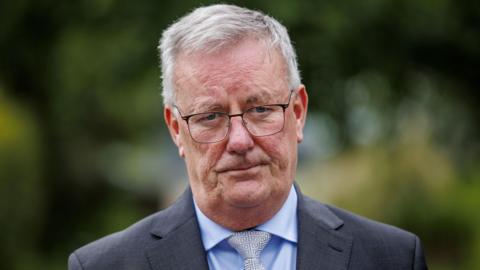 Mike Nesbitt is wearing a grey blazer, blue shirt and grey tie. He has black glasses on his face. He has short grey hair. The background is blurry. 