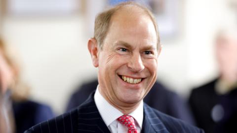 Prince Edward is wearing a navy pin-stripe suit, with a patterned red tie. The background is blurred.