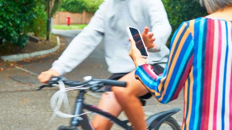 A man in a grey hoodie cycles past a woman using her phone, and reaches to snatch it out of her hand