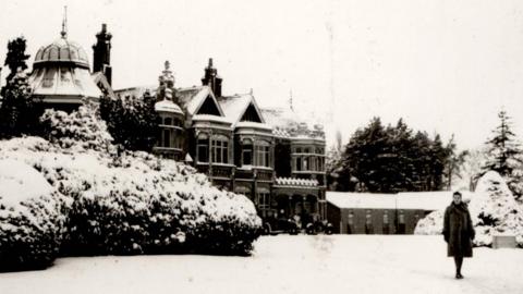 A woman, standing on white snow, wearing a coat, standing by an old Bletchley Park building. Tere is snow on the ground, in the trees and on top of the building. The picture is in black and white.
