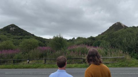 Two people with their backs to the camera look at two pointy peaks that look like small hills.They are actually mine waste from Cornish china clay production. 