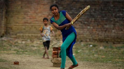 A woman, wearing a blue and green outfit, swings a cricket bat on February 27, 2024 in Ranchi, India. Behind her is a makeshift wicket made of bricks and stones. A young boy wearing a T-shirt and shorts is visible in the background.