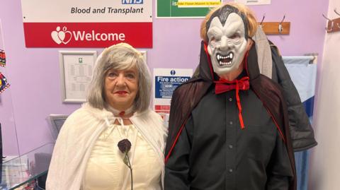 Carol is on the left, dressed in fancy-dress - she is wearing a mock white wedding dress with a thin white cloak, and is holding a dark purple rose up to her chest. On her right is her husband dressed in a black shirt with red bowtie and a black and red hood, wearing a latex mask of Dracula. In the background signage for the blood donor centre can be seen. 