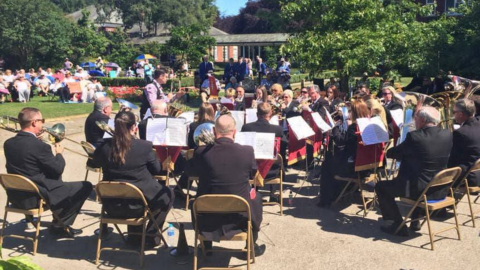 A full brass band are playing for people lounging in a field. There are about two dozen people wearing black suits sitting in a semi-circle around the conductor. There are playing instruments like trumpets and French horns, while looking at sheet music on stands in front of them.