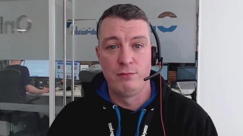 A still from a video call of Cas Paton sitting in an office, wearing a headset and looking down the lens. He has short brown hair and is wearing a black hoody. Behind him is a glass screen and beyond the screen are the backs of other workers sitting at their computers.