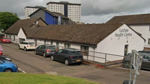 Lochee Health Centre which is a long, white, siongle-storey building with a slate rood. There are flats and houses areound it and cars in a car park at the front and side.
