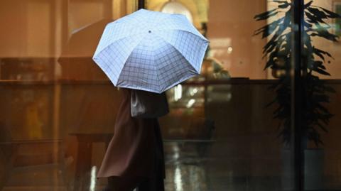 A figure holds an umbrella above their head as they walk past a large window. Their face cannot be seen beneath the umbrella.