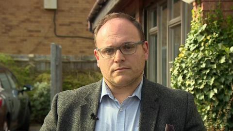 Dr Jamie Green is unsmiling while photographed outside a building. He is wearing glasses and a grey blazer and light blue shirt stands. You can see a car behind him and some ivy on the side of a building. 