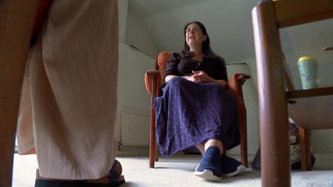 The image shows Emma Eveleigh, who is a client of the Haye Mill counselling service. She is sitting in a chair in the counselling room facing her therapist. Emma is wearing a purple top with a purple flowery skirt and blue trainers.