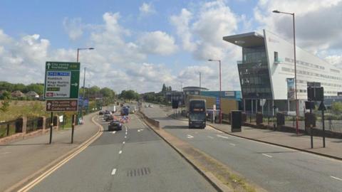 A dual carriageway with cars on one sign, a green and white sign for directions and a bus on the other side. A metal railing goes down the central reservation.