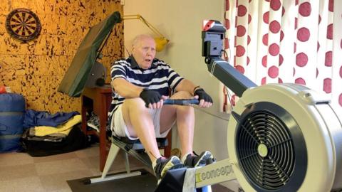 A man wearing a t-shirt with stripes and shorts on, sits on a rowing machine in a home gym. 