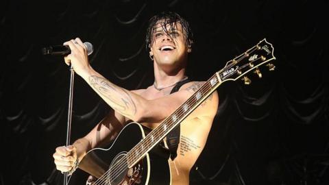 Yungblud is topless on stage. He's wearing black leather trousers and is holding a microphone looking out into the crowd. Behind him is a giant festival type stage with two screens flanking the sides with him on it.