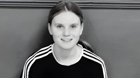 Black and white picture of Holly. She is sitting wiht her back against a wall and is smiling at the camera