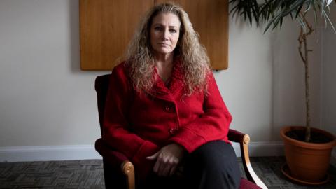 Marlean Ames ints in a chair weraring a red blazer  and black pants with her hair falling over her shoulders