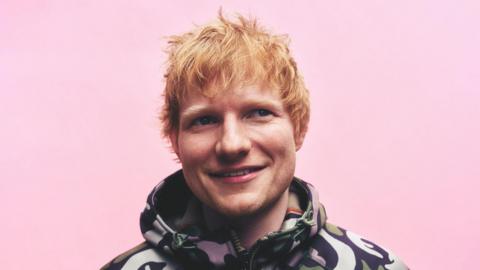 A press photograph of Ed Sheeran. He is wearing a jacket with a pattern on. He has short red hair and is in front of a pink background.