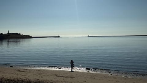 A woman standing on a beach looking out to the sea. Two piers can be seen in the distance.
