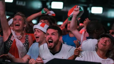 A fan in a white shirt and St George's Cross headband screams with clenched fists at Boxpark Croydon. Other fans around him shout and wave flags.