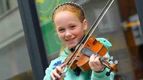 a red-headed girl in a green hoodie playing the fiddle and smiling at the camera