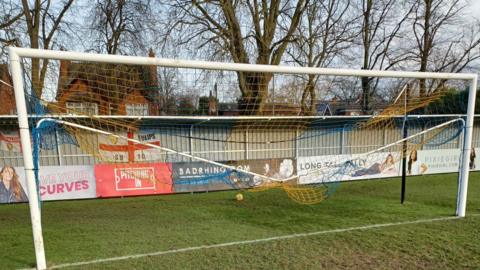 A white goal with orange and blue netting. One of the posts has been bent 