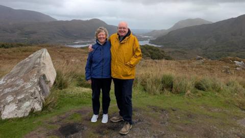 Older woman with grey hair in blue jacket and an older man in a yellow jacket smiling together with a rural backdrop including a lake and hills.