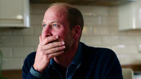 Prince William holds a hand over his mouth, he's sitting in a white tiled kitchen and wearing a navy blue jumper