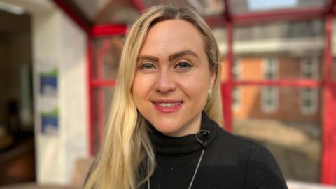 Rhi has long, blonde hair and is wearing a black roll-neck top. She is stood in a room with red-framed windows behind her, which are out of focus.