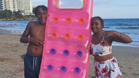 Ameiya (R) and Ricardo Del Brocco (L) on holiday. They are either side of an pink inflatable. They are on a beach and smiling.  Ameiya has short dark hair and wears a two piece costume of white with a floral design. Ricardo has short dark hair in braids and wears a blue pair of shorts. 