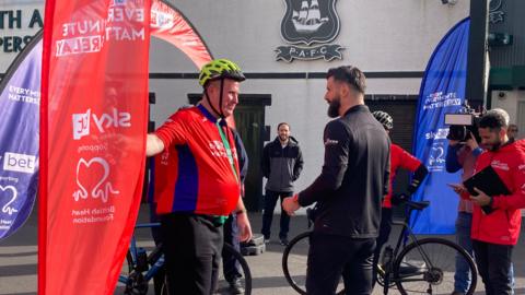 Plymouth Argyle manager Miron Muslic is pictured with a Plymouth Argyle fan who is starting the relay off from Home Park. There are red and blue flags around the side and two bicycles pictured in the background.