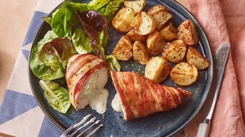 A blue plate of potatoes salad and a halved chicken breast wrapped in bacon with a creamy middle, with a pink linen cloth