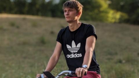 A young man wearing an adidas top cycles on a bike, with a field in the background