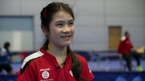 Alyssa smiles looking past the camera. She is wearing a red England top and there are table tennis tables in in the blurred background.