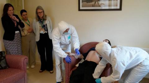 Devon and Cornwall Police and Crime Commissioner Alison Hernandez (left) watches alongside two other women - one of whom is Plymouth Marjon University vice-chancellor Professor Claire Taylor - as two students search a dummy during an exercise on a forensics course at the university. The exercise is taking place in a living room with a picture on the wall and two red sofas.