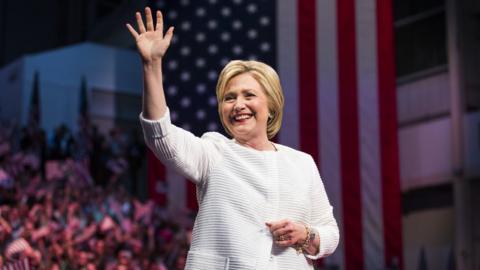 Hillary Clinton waving to crowds in front of the US flag