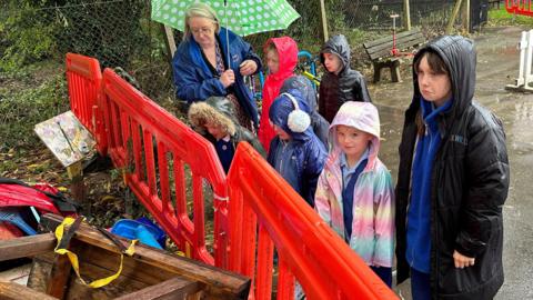 Children wearing coats and hoods stand in the rain looking sadly over an orange safety barrier at destroyed play equipment