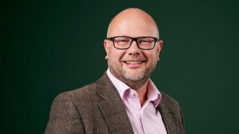 A bald man with a beard is smiling at the camera, he is wearing black glasses. He is wearing a brown tweed jacket and a pink shirt