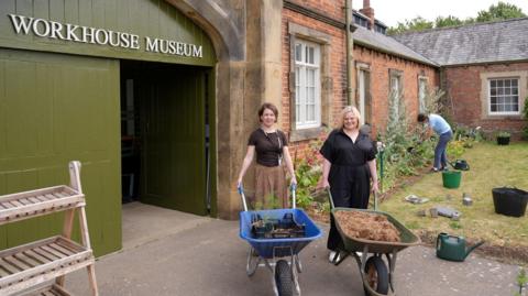 Museum director Alexa Vernon Museum and curator Dr Laura Allan stand with wheelbarrows outside the entrance to a workhouse museum.