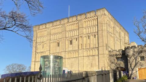 A Norman castle is shown under blue skies with a circular glass elevator outside it and iron railings in the foreground.