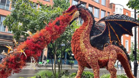 A red dragon breathing fire on a busy urban street. Both the dragon and the trail of fire are made out of red flowers.