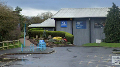 Google Street View of Two Riversmeet leisure centre viewed from the car park on a grey rainy day. The grey warehouse-style building has a pitched roof. A curved hedge and a rockery area planted with shrubs separates the car park from the entrance.