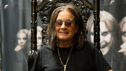 Ozzy Osbourne sits on a dark wood, high-backed chair with ornate designs on it. He is wearing round sunglasses, and is smiling, while also wearing a black T-shirt and a silver pendant chain. Behind him there are images of him on canvas with different facial portraits.
