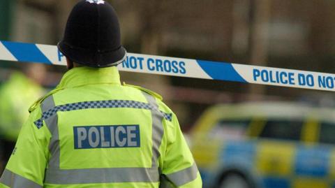 The back of a police officer in high visibility yellow jacket with Police written on the back, standing in front of crime scene tape 