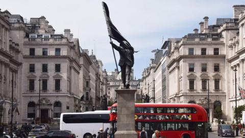 Statue of a man holding a flag in central London. A London bus and people walking around in the background