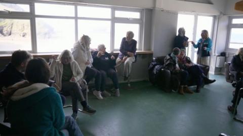 A picture of a group of people sat in a large room located on beach. Some people are stood up, with a majority of people sat down.