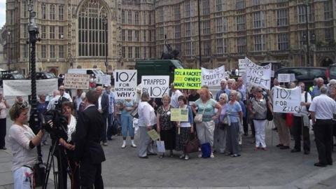 A group of park home residents are at Westminster in 2017 holding protest signs about a 10% commission fee. Several people hold placards reading “10% for nothing why?”, “Trapped by 10%” and “10% equity theft”, while a television crew films nearby in front of the building.