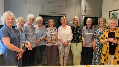 A group of 10 women in their 70s and 80s standing in a large kitchen. They are all smiling.