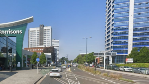 Street view of Hagley road in Birmingham. On the left a Morrisons store can be seen with and office building beyond it, and another one to the left. Cars can be seen crossing the tracks of the West Midlands Metro network, while on the opposite carriageway, vehicles queue for the Five Ways interchange.